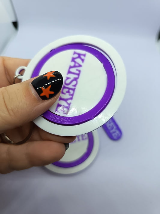 Llavero KATSEYE con logo giratorio morado, fotografía 2 del producto.